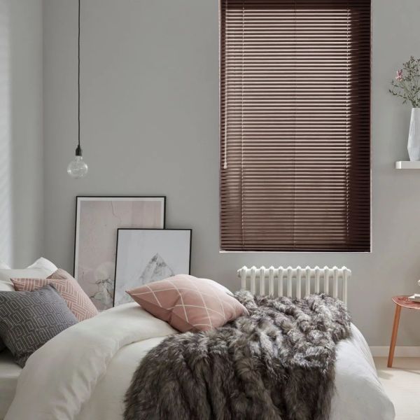 Brown horizontal slatted blinds, lowered and closed, slightly filtering daylight; bedroom setting with grey walls, white bed topped by pink and grey pillows and a grey faux-fur throw, radiator below.