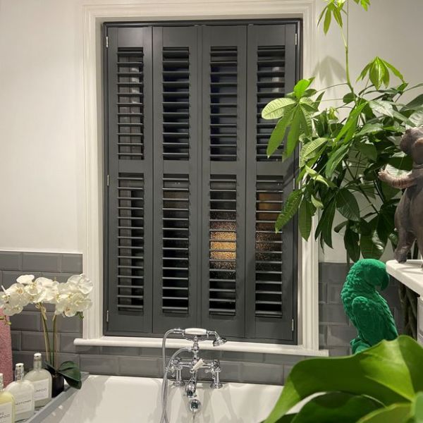 Dark grey four-panel interior louvered shutters covering window, panels closed with slats angled to filter light; set above a bathtub with chrome faucet, grey subway tiles, orchids and houseplants nearby.