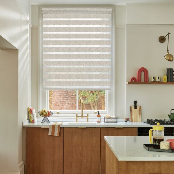 Beige dual-layer zebra roller blind, lowered covering most of the window, alternating opaque and sheer horizontal stripes filtering daylight; modern kitchen sink area with brass faucet, wooden cabinets, marble countertops.