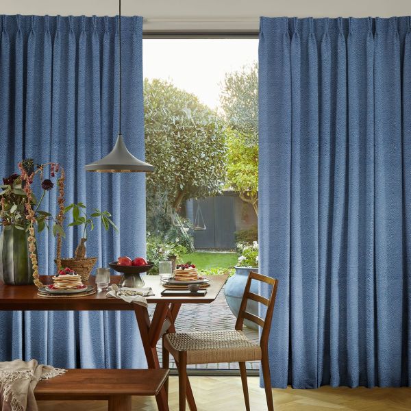 Pleated floor-length blue curtains, parted at center to frame a glass patio door, filter soft daylight into a cozy dining area with table and leafy garden beyond.