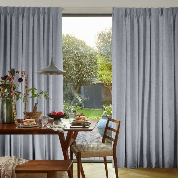 Gray pleated floor-length curtains, parted center to reveal glass door, soft textured fabric. They frame a sunlit dining nook with wooden table, pendant lamp, and a green backyard with trees.