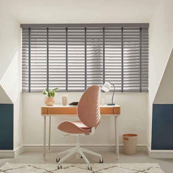 Grey horizontal Venetian blinds, fully lowered with dark tapes, filtering daylight and casting stripes across a small attic office with a wooden desk, pink swivel chair, lamp, plant.