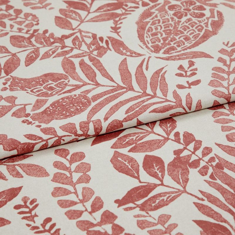 Cream fabric printed with terracotta botanical and pomegranate motifs, folded and overlapped to show pattern repeat, close-up on a flat surface under soft, even studio lighting.