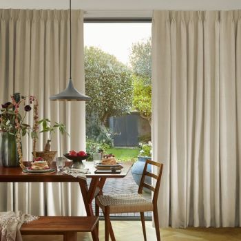 Full-length beige pleated curtains framing a glass sliding door, partly drawn to reveal a sunlit garden; soft daylight filters into a cozy dining area with wooden table and chairs.