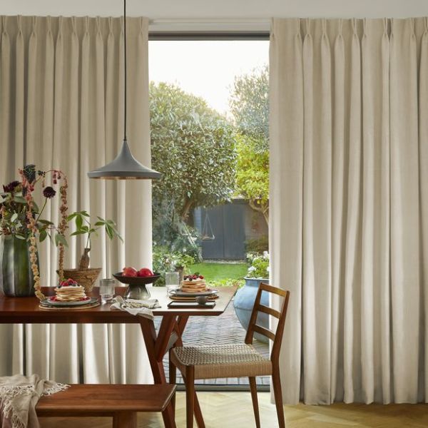 Full-length beige pleated curtains framing a glass sliding door, partly drawn to reveal a sunlit garden; soft daylight filters into a cozy dining area with wooden table and chairs.