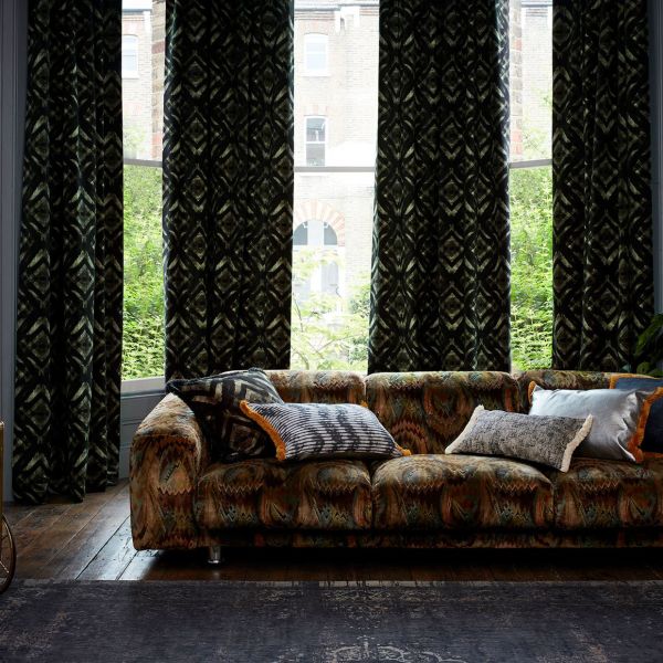 Heavy dark green patterned curtains, mostly drawn with central gaps, filtering daylight through tall bay windows; in front, a low patterned sofa with cushions on wooden floor, brick view beyond.