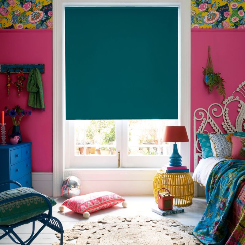 Teal roller blind, fully lowered and blocking most light, covering a white-framed sash window; colorful bohemian bedroom with hot-pink walls, patterned wallpaper border, ornate white bed and blue dresser.