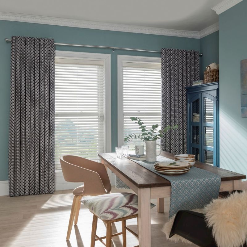 Patterned drapes on a rod flank two tall windows; lowered white horizontal blinds filter daylight in a sunlit teal dining room with wooden table, chairs, table runner and a blue cabinet.
