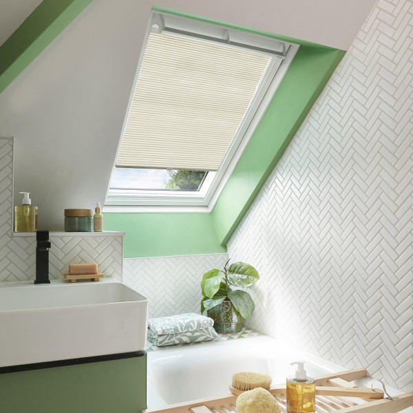 Cream pleated skylight blind, lowered halfway and filtering daylight, in a bright sloped-ceiling bathroom with green trim, white herringbone tiles, sink, bathtub and potted plant.
