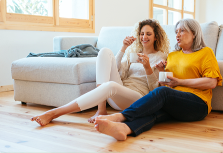Two barefoot women sitting next to each other against a couch on a hardwood floor, they're holding mugs of tea.
