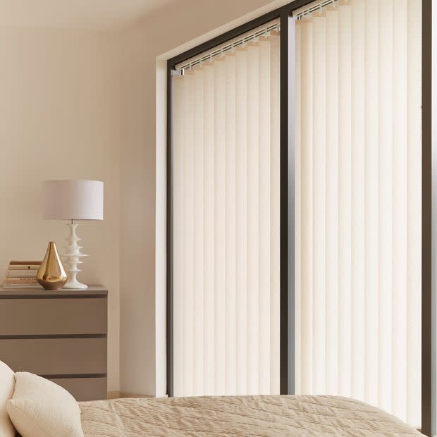 Vertical fabric vertical blinds covering two tall sliding glass doors, closed and softly diffusing daylight; beige bedroom with quilted bed, bedside dresser, lamp and decorative gold vase.