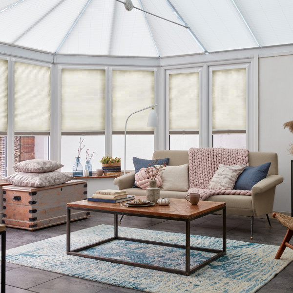 Cellular pleated blinds, cream-colored and lowered on bay conservatory windows, filtering soft daylight; bright sunroom with beige sofa draped in a chunky pink throw, wooden coffee table and rug.