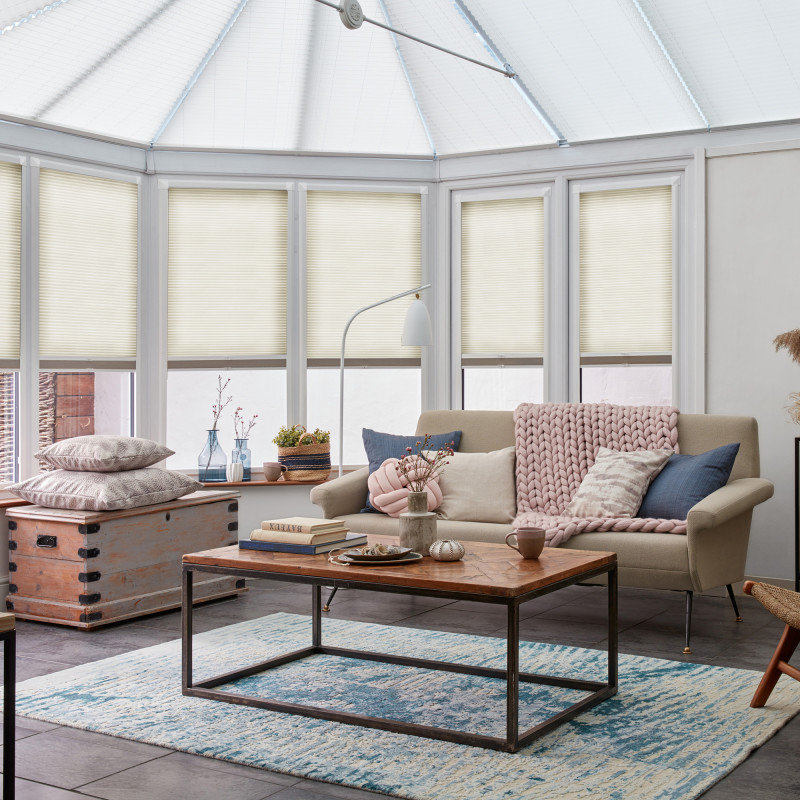 Cellular pleated blinds, cream-colored and lowered on bay conservatory windows, filtering soft daylight; bright sunroom with beige sofa draped in a chunky pink throw, wooden coffee table and rug.