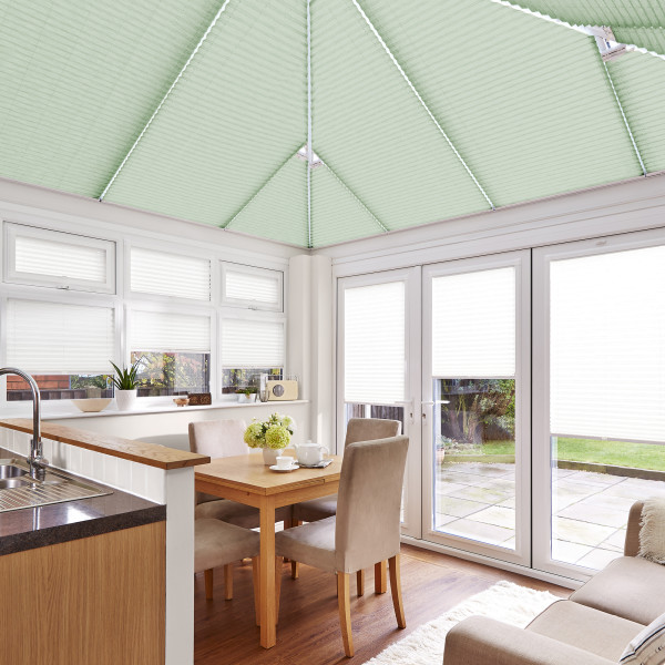 Mint-green pleated roof blinds spanning the pitched conservatory ceiling, closed and diffusing light; matching white cellular blinds on windows and doors; small dining area and kitchen beside a sunlit patio.