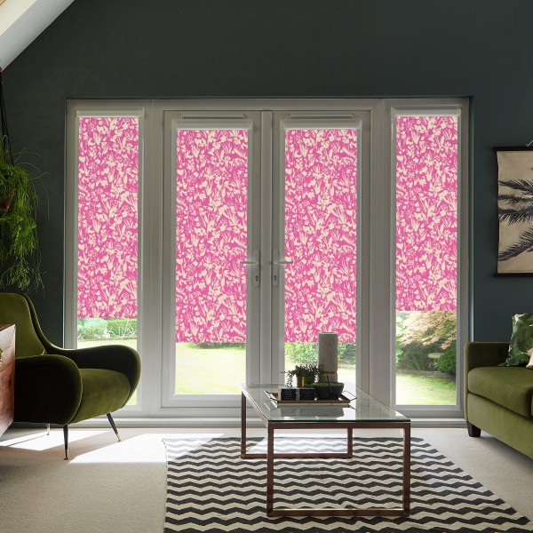 Four tall window panels are fitted with bright pink floral roller blinds, blocking light in a modern living room with green armchairs, glass coffee table, chevron rug, and garden beyond.