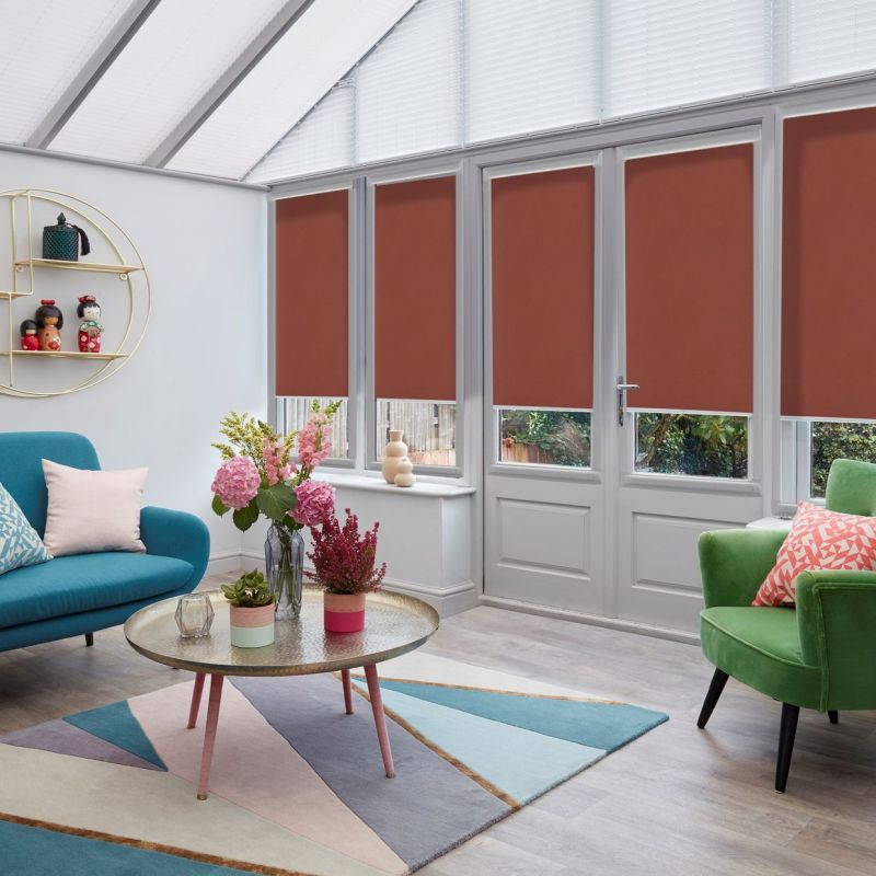 Brown roller blinds covering four tall conservatory windows and door, lowered mid-height to block light; modern sunroom with glass roof, teal sofa, green armchair, floral centerpiece on round coffee table.