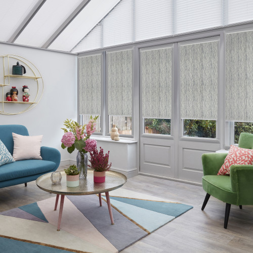 Light-grey textured fabric roller blinds lowered over the upper two-thirds of tall windows and French doors, filtering daylight into a bright conservatory-style living room with colorful sofas and coffee table.