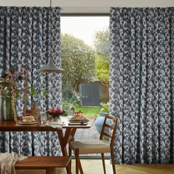 Full-length pleated geometric blue-gray curtains, drawn to sides and framing a central sliding glass door, filtering soft daylight; dining room with wooden table set for breakfast, pendant lamp, leafy backyard.