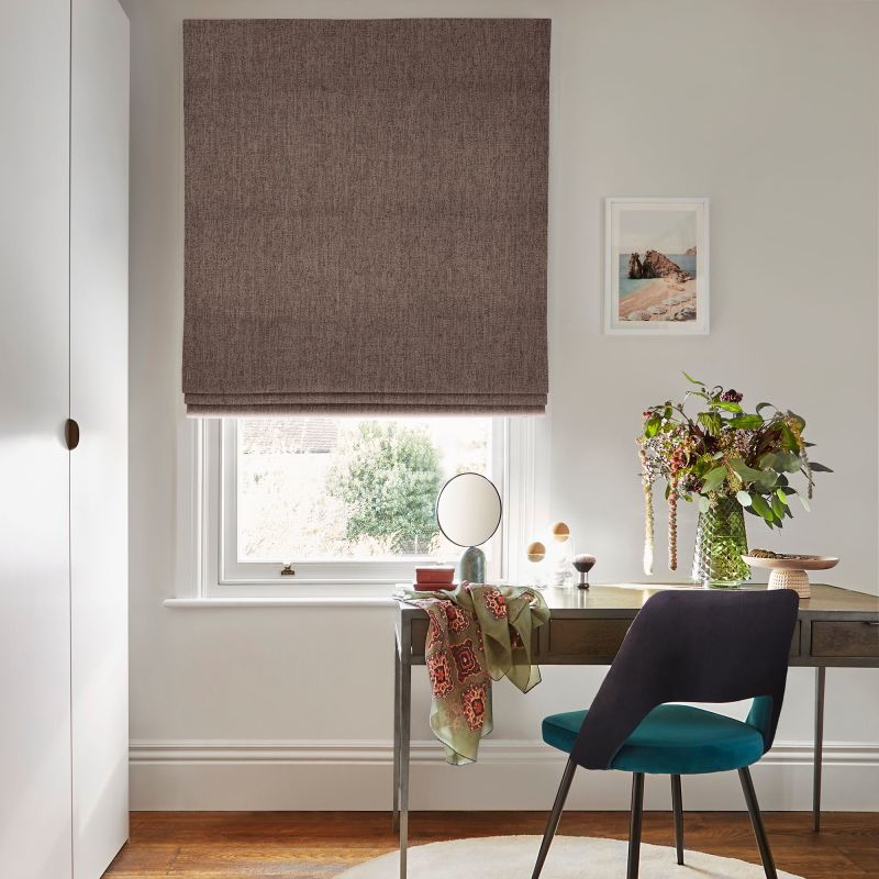Brown fabric Roman blind, fully lowered with a narrow light gap, textured finish; filtering daylight over a wooden desk with chair, mirror and vase of flowers in a sunlit home office.