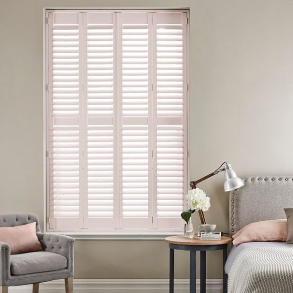 Four-panel pale-pink plantation shutters, full-height with adjustable louvers, closed and softly diffusing daylight; bedroom corner with upholstered bed and headboard, bedside table lamp and vase, tufted armchair.