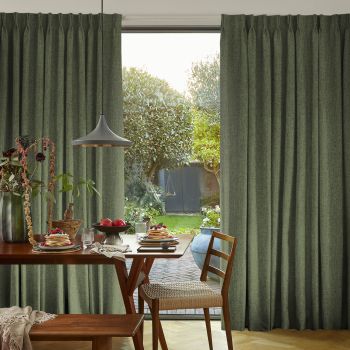 Floor-length, pleated olive-green curtains, drawn open at the center, framing a sliding glass door; they reveal a sunlit dining corner with a wooden table set for breakfast and a leafy backyard.