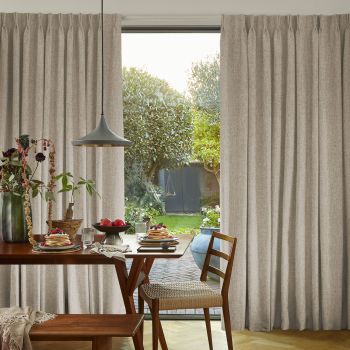 Beige floor-length pleated curtains, hung and parted at center revealing a glass door, filtering soft daylight onto a wooden dining table set for breakfast, with a green garden visible beyond.