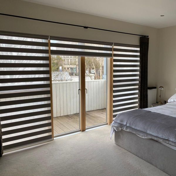 Zebra roller blinds with alternating dark and translucent horizontal stripes, partially lowered; curtains drawn right. Central sliding doors reveal a small balcony in a lit bedroom with grey bedding.