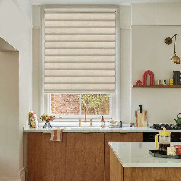 Layered beige zebra roller blind, lowered and filtering soft daylight across the window; modern kitchen context with wooden cabinets, white marble countertops, brass sink faucet and a brick-and-greenery view.