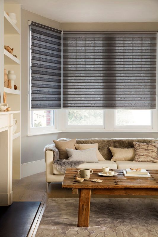 Layered horizontal roller blinds covering a bay window, dark grey wood-grain bands, lowered and filtering daylight; above a neutral living room with beige sofa, fur throw and rustic coffee table.