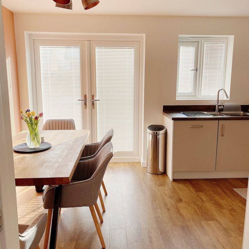 White horizontal Venetian blinds cover the French doors and kitchen window, fully closed filtering soft daylight into a dining-kitchen with wooden table, upholstered chairs, sink, vase of flowers.