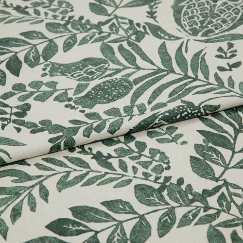 Botanical-printed cotton fabric, folded across the center revealing a layered edge, green leaf-and-fruit motifs on an off-white ground; close-up textile sample on a neutral surface under soft studio light.
