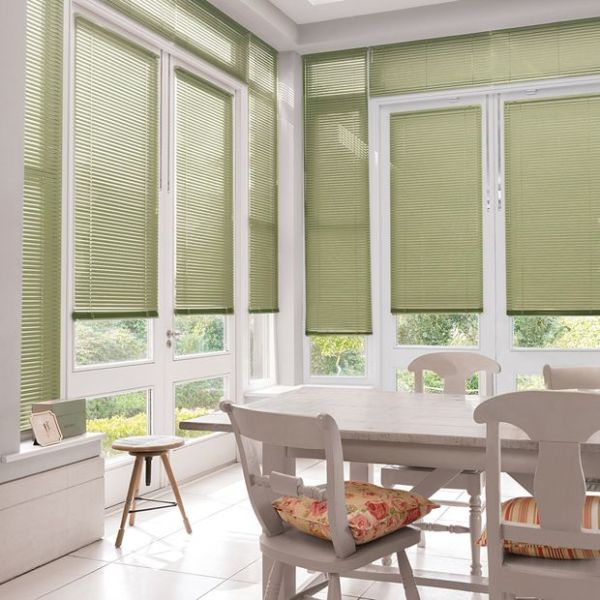 Green pleated window blinds, fully lowered across tall French doors and surrounding windows, lightly filtering sunlight; dining room with white wooden table and chairs, floral cushions, tile floor, garden view.