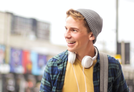 A smiling teen boy wearing a knitted gray hat and a yellow t-shirt under a blue and green flannel shirt with headphones around his kneck.