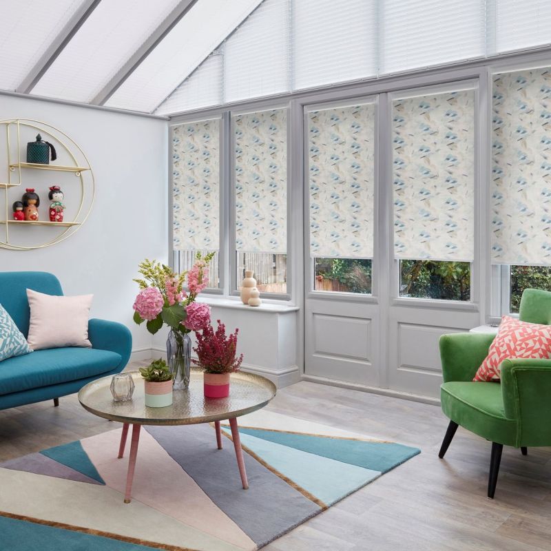 Floral-patterned roller blinds cover the lower panes of tall conservatory windows and doors, partially lowered to filter daylight; bright seating (teal sofa, green armchair), round coffee table and flowers.