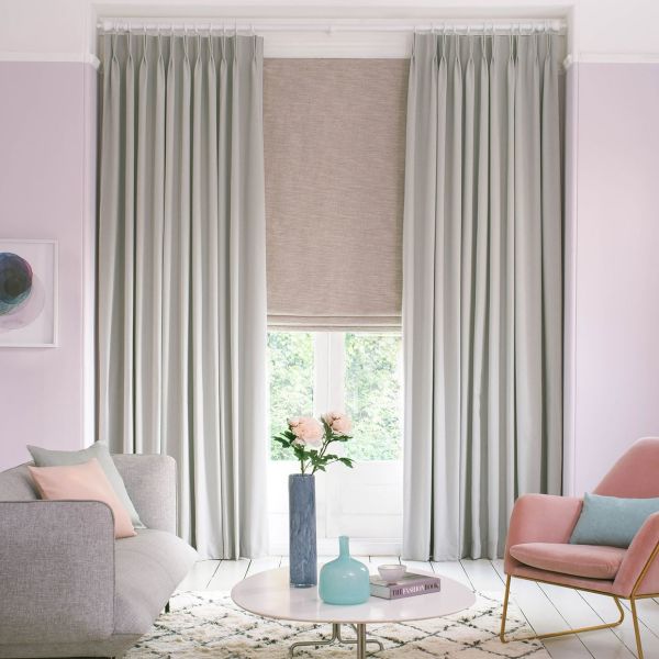 Floor-length pleated grey curtains frame a lowered beige Roman blind; drawn to the sides while the blind filters light, revealing a leafy view in a softly lit pastel living room.
THE FASHION BOOK