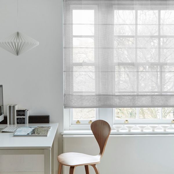 Sheer fabric Roman blind lowered across a multi-pane window, softly filtering daylight; minimalist home office with white desk, wooden chair, paper pendant lamp, and faint leafless-tree view outside.