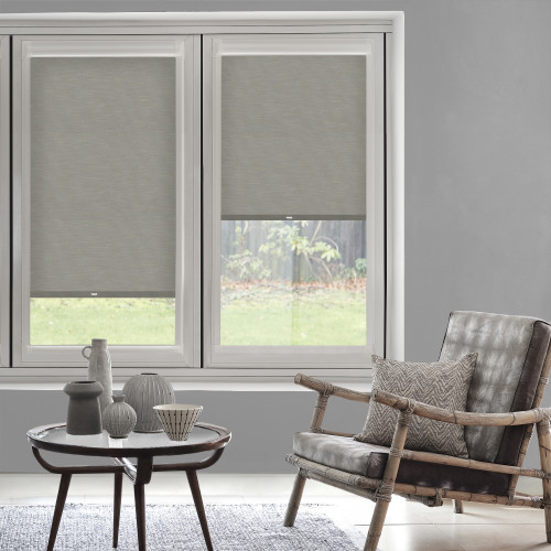 Grey fabric roller blinds on two windows, partially lowered with pull tabs, filtering daylight; neutral living room with wooden-frame armchair, patterned cushion, round coffee table, backyard lawn visible.