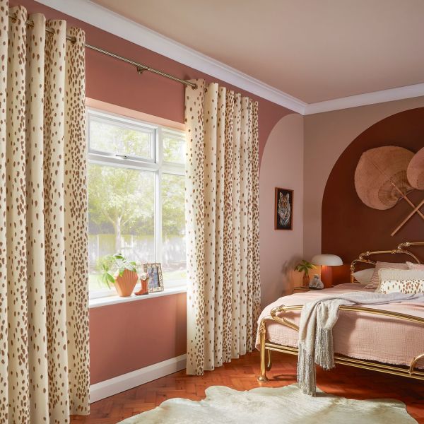 Cream grommet curtains with brown spots, hung on a brass rod, parted to frame and softly filter daylight through the window; warm pink bedroom with a brass bed and plants.