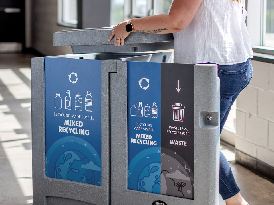 indoor grey plastic trash and recycling bin with removable lid and large body signs in the entrance of a hockey arena showing a woman servicing the bin