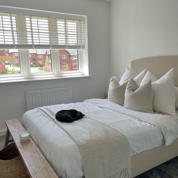 White slatted blinds, partially lowered and filtering daylight into a bright bedroom with a beige upholstered bed, white bedding, neutral pillows, a curled black cat, and red‑brick houses outside.