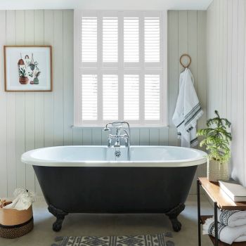 White plantation shutters covering a tall window, slats closed and filtering bright daylight; set above a black clawfoot bathtub in a light‑paneled, softly lit bathroom with towel and potted plant.