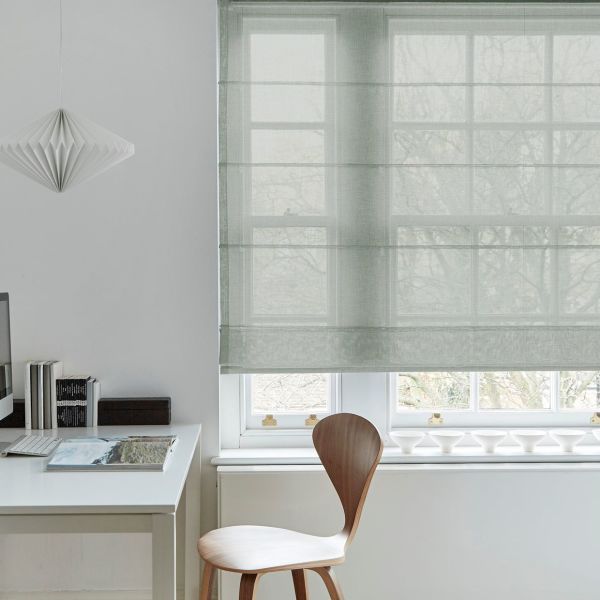 Sheer pale-gray Roman blind lowered over a multi-pane window, softly filtering daylight and revealing tree silhouettes; a minimalist home office with a white desk, wooden chair and pendant lamp.