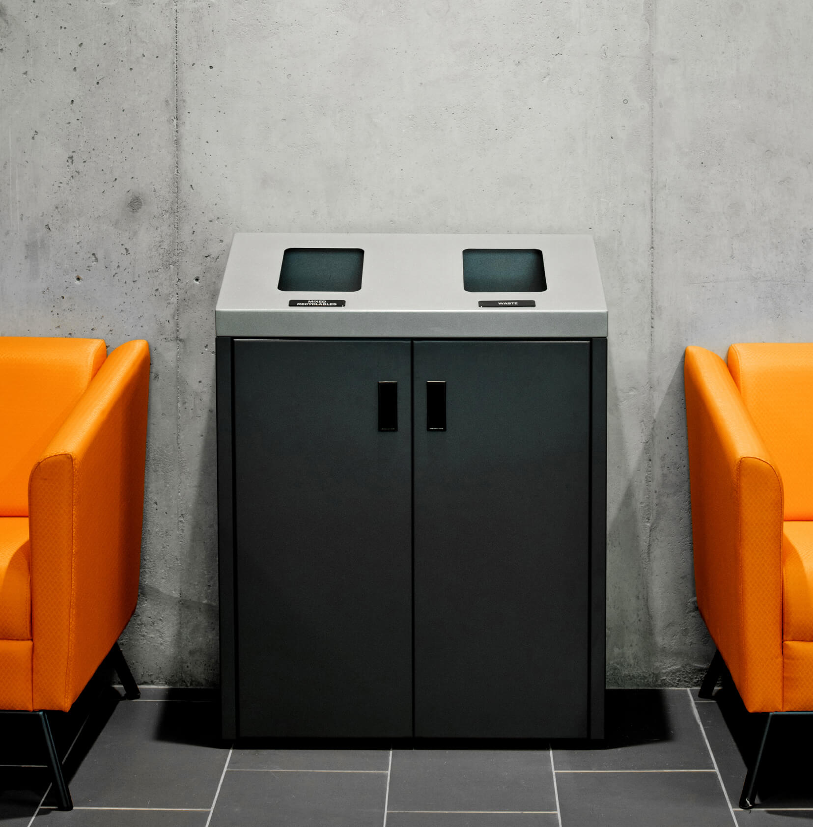 double waste and recycling bin against a concrete wall between two orange chairs