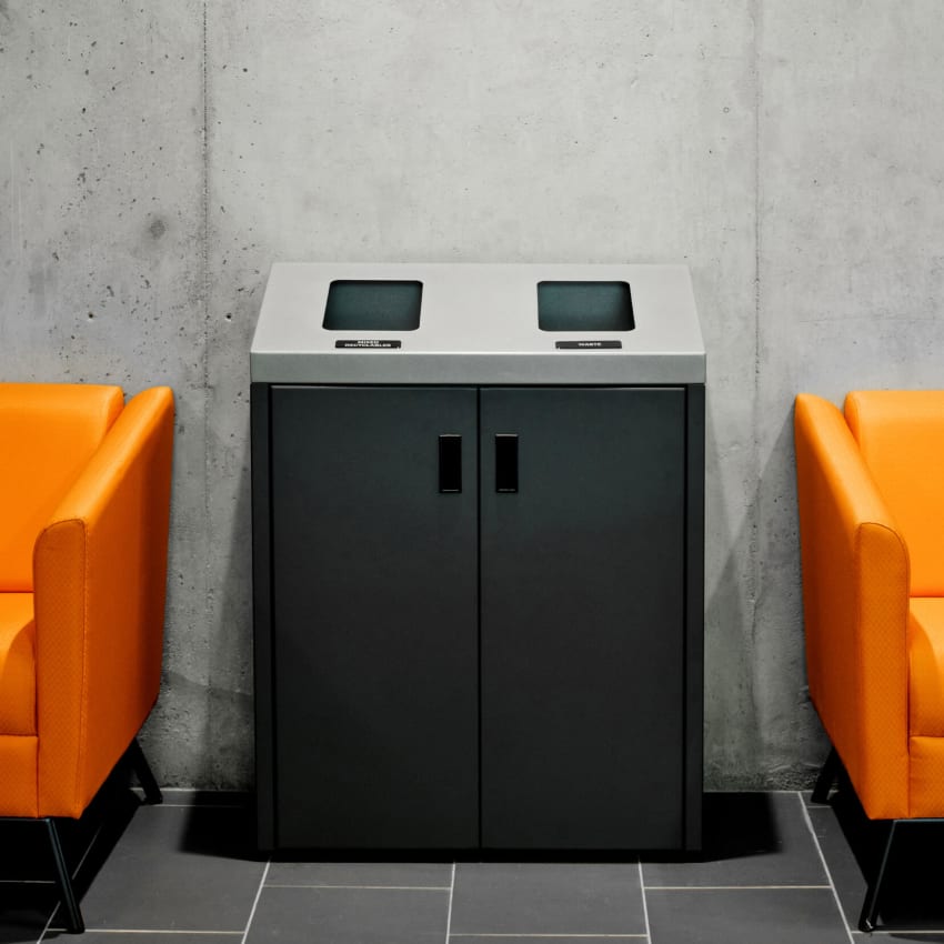 double waste and recycling bin against a concrete wall between two orange chairs