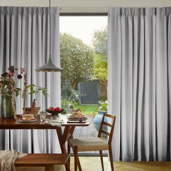 Grey, floor-length pleated curtains framing a sliding glass door, slightly parted to admit soft daylight — wooden dining table set for breakfast with pancakes, pendant lamp, and garden view.
