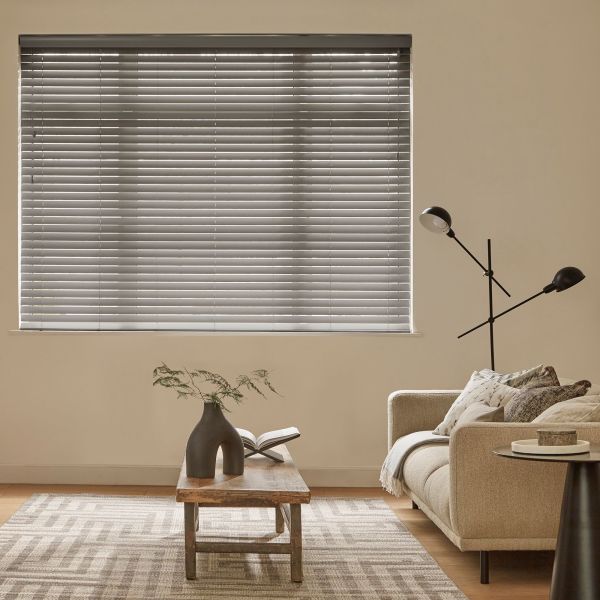Horizontal venetian blinds covering a large wall window, slats mostly closed and filtering soft daylight; beige living room with sofa, wooden coffee table, patterned rug and black floor lamp.
