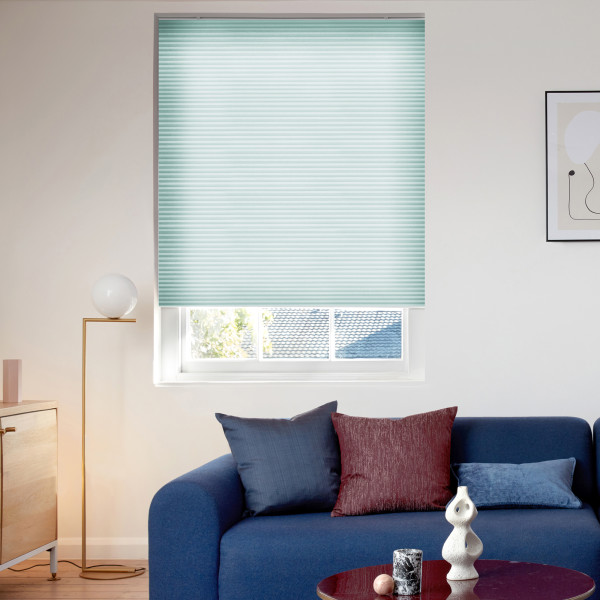 Light teal cellular blind, mostly lowered across a recessed window, softly filtering daylight; modern living room with a blue sofa, colored cushions, brass floor lamp and minimal art.