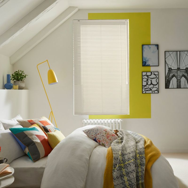 White horizontal blinds covering a window, closed and filtering daylight; in an attic bedroom with sloped ceiling, yellow wall accent, bed with colorful pillows and a yellow floor lamp.