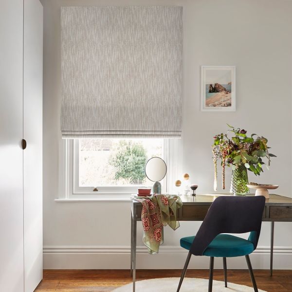 Textured beige Roman blind, lowered with a fold at the bottom, filtering daylight; above a window over a wooden desk with teal chair, vase of flowers and framed seaside print.