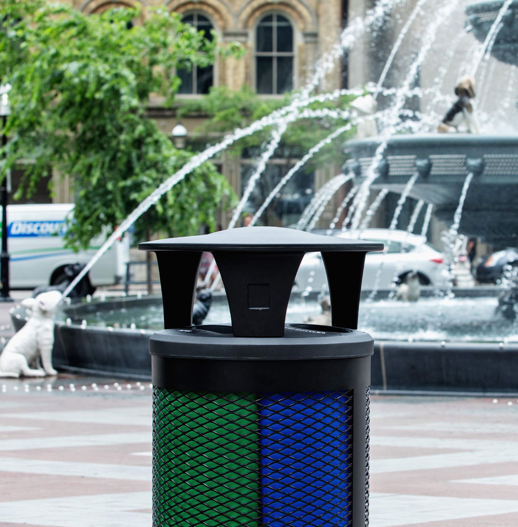 black metal outdoor waste and recyclables container with a plastic canopy lid at a city park with a water fountain